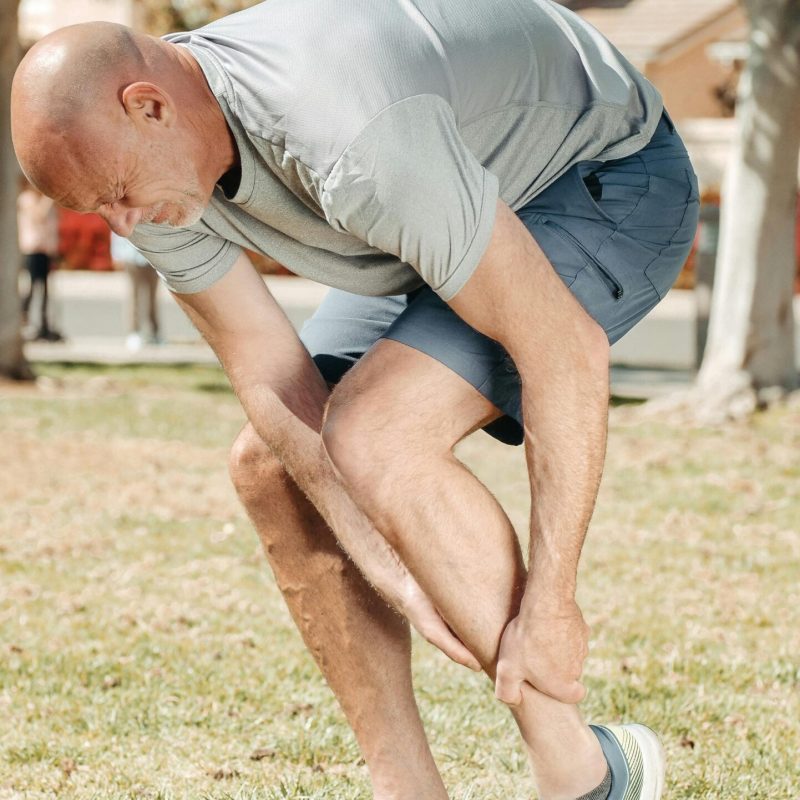 Man holding his injured ankle outdoors in a park, highlighting running-related pain or sport injury.